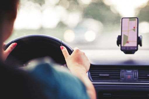 A driver holds a steering wheel with a smartphone mounted on the dashboard.