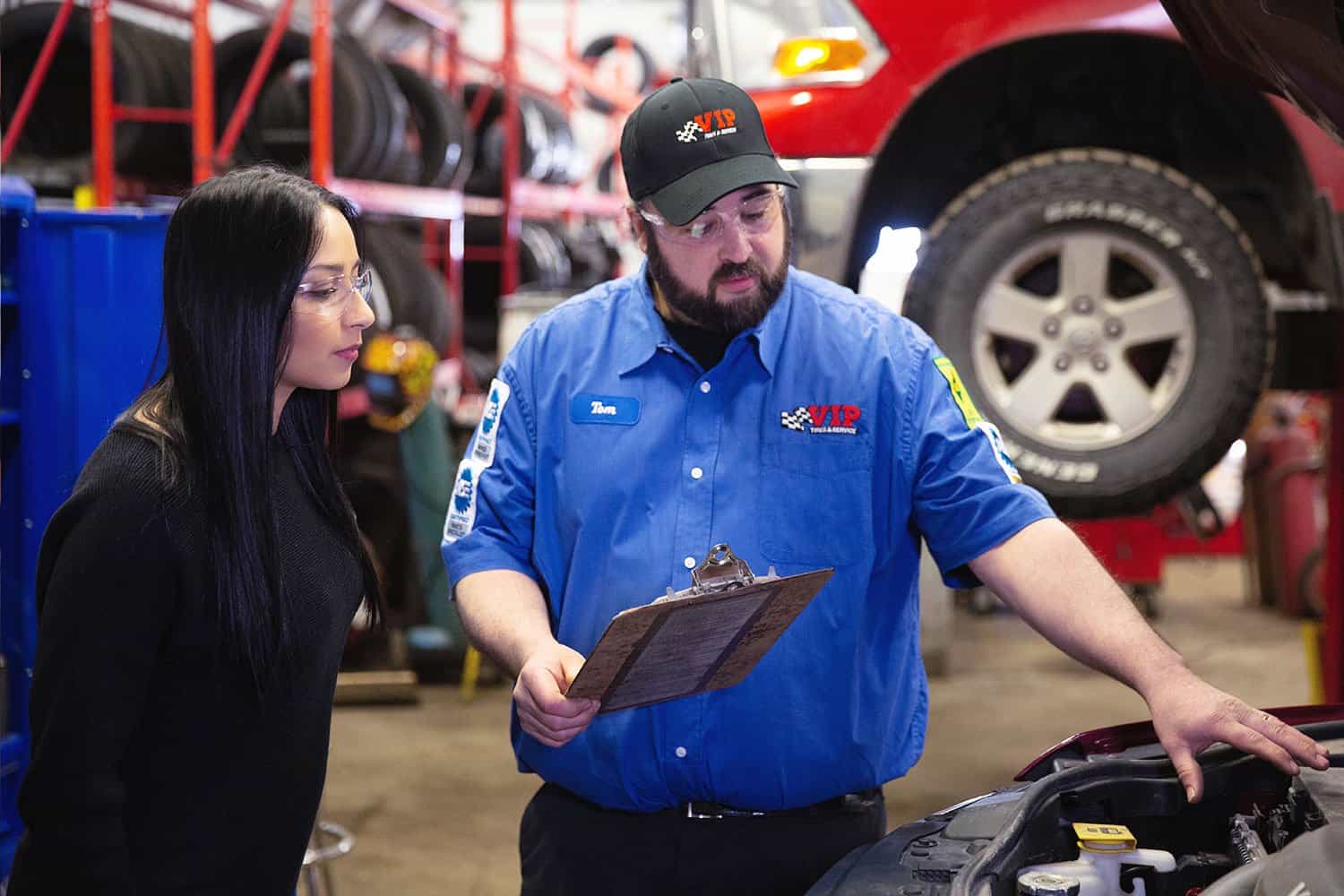 The image captures a professional interaction in an auto repair shop, featuring a mechanic and a customer. The mechanic, wearing a blue uniform with a VIP Tires & Service logo, is explaining the details on a clipboard to the female customer. They are standing in a well-equipped garage with a car raised on a lift in the background. This scene portrays a typical customer service situation in the automotive industry, emphasizing the importance of clear communication and expert advice in customer interactions.