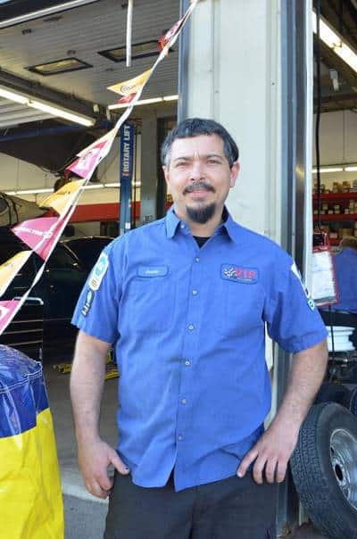 Man in blue work shirt standing in front of a vehicle in a garage.