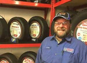 Man in blue uniform and cap standing next to stacked tires on red shelving.
