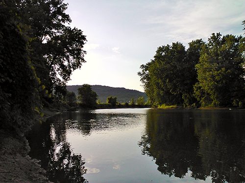 A calm river reflects trees and sky, with forested hills visible in the background.