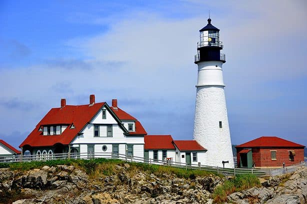 A white lighthouse with a black top stands beside a red-roofed keeper's house on a rocky coastal.