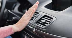 The image shows a close-up view inside a car, focusing on a person's hand held near the air conditioning vent to check the airflow. The dashboard is visible in the background, highlighting modern car interior elements. This visual is typically used to illustrate issues related to car air conditioning systems, particularly when the A/C is not blowing cold air, indicating potential problems like low refrigerant levels, a faulty compressor, or blocked air filters.