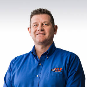 A man wearing a blue button-up shirt with an embroidered logo smiles at the camera.