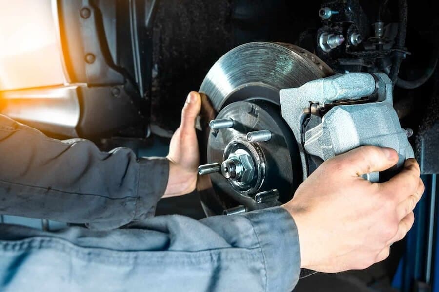 A close-up image of a mechanic's hands installing a brake caliper onto the disc brake assembly of a vehicle. The mechanic is wearing a grey uniform and is working within the wheel well, illustrating a car maintenance or repair scene.