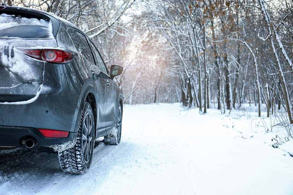 The rear view of a dark-colored SUV parked on a snowy forest road, its tires covered in fresh snow, depicting a cold winter day. The vehicle’s brake lights cast a warm glow on the snow, contrasting with the cool tones of the surrounding winter landscape.