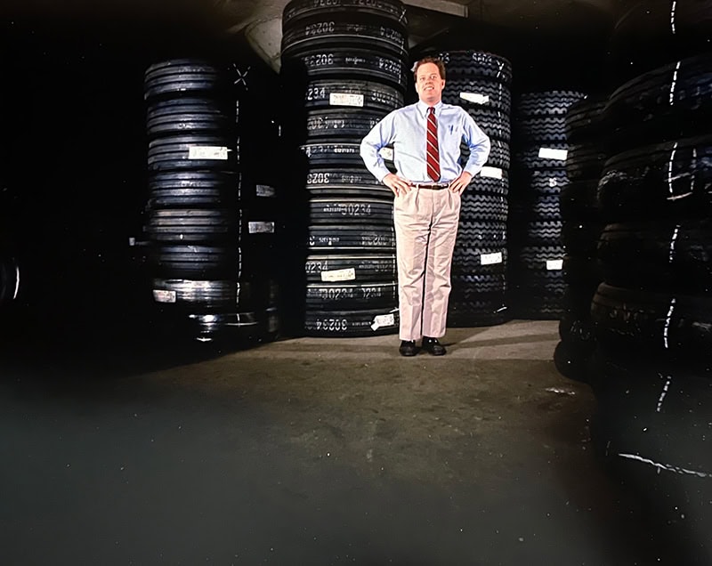 Man in business attire stands among stacked tires in a warehouse.
