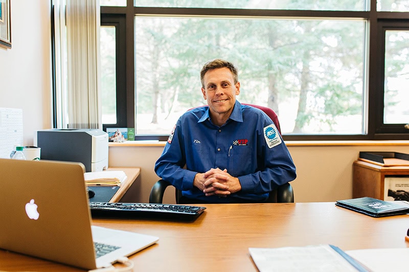 Man in blue work shirt sitting at desk with laptop and keyboard in an office with large windows.