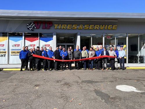 Group of people holding a red ribbon in front of a tire service shop during a ribbon-cutting.