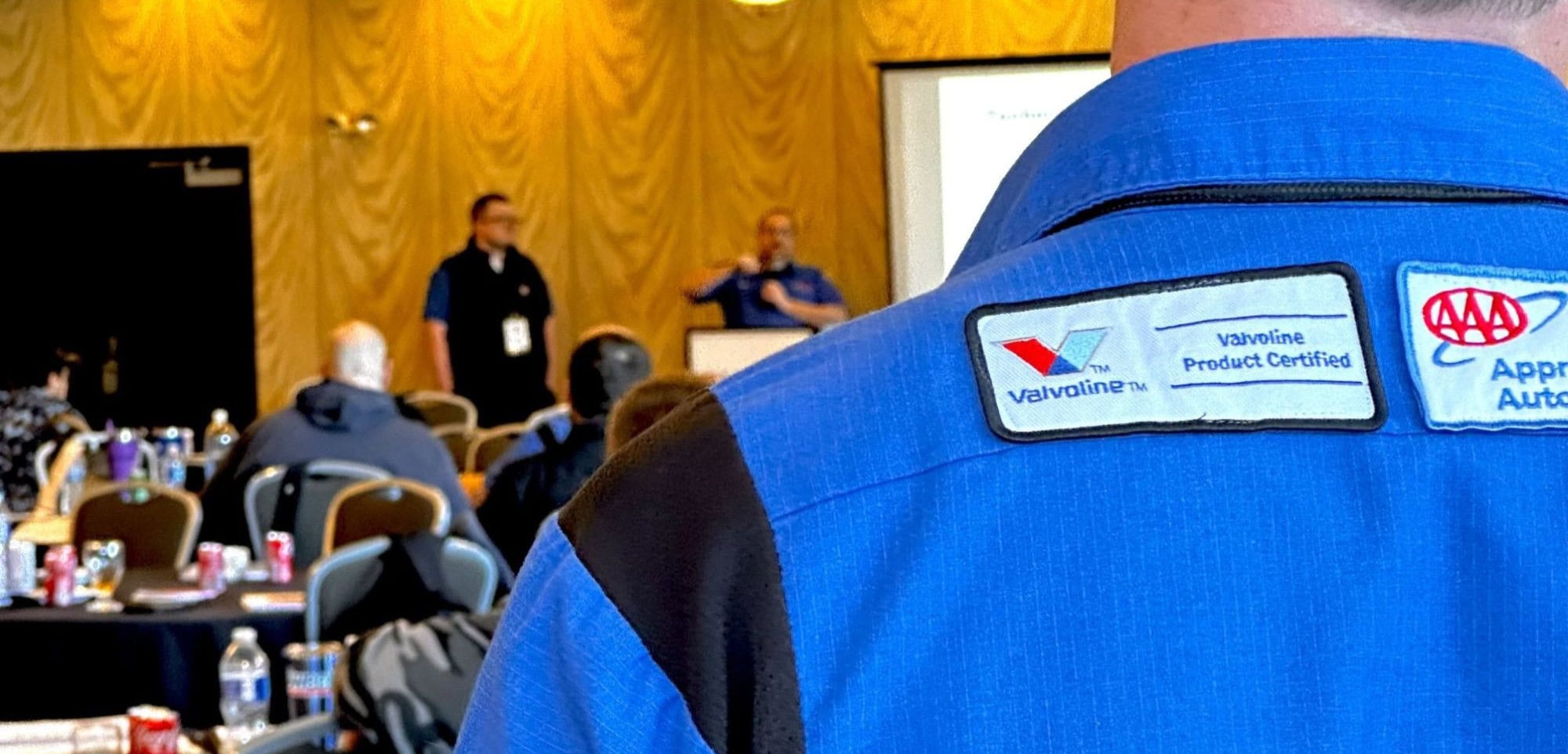 A blue Valvoline-branded jacket displays AAA and product certification logos at an indoor event.