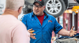 Mechanic in blue work uniform and cap gestures while speaking in a garage.