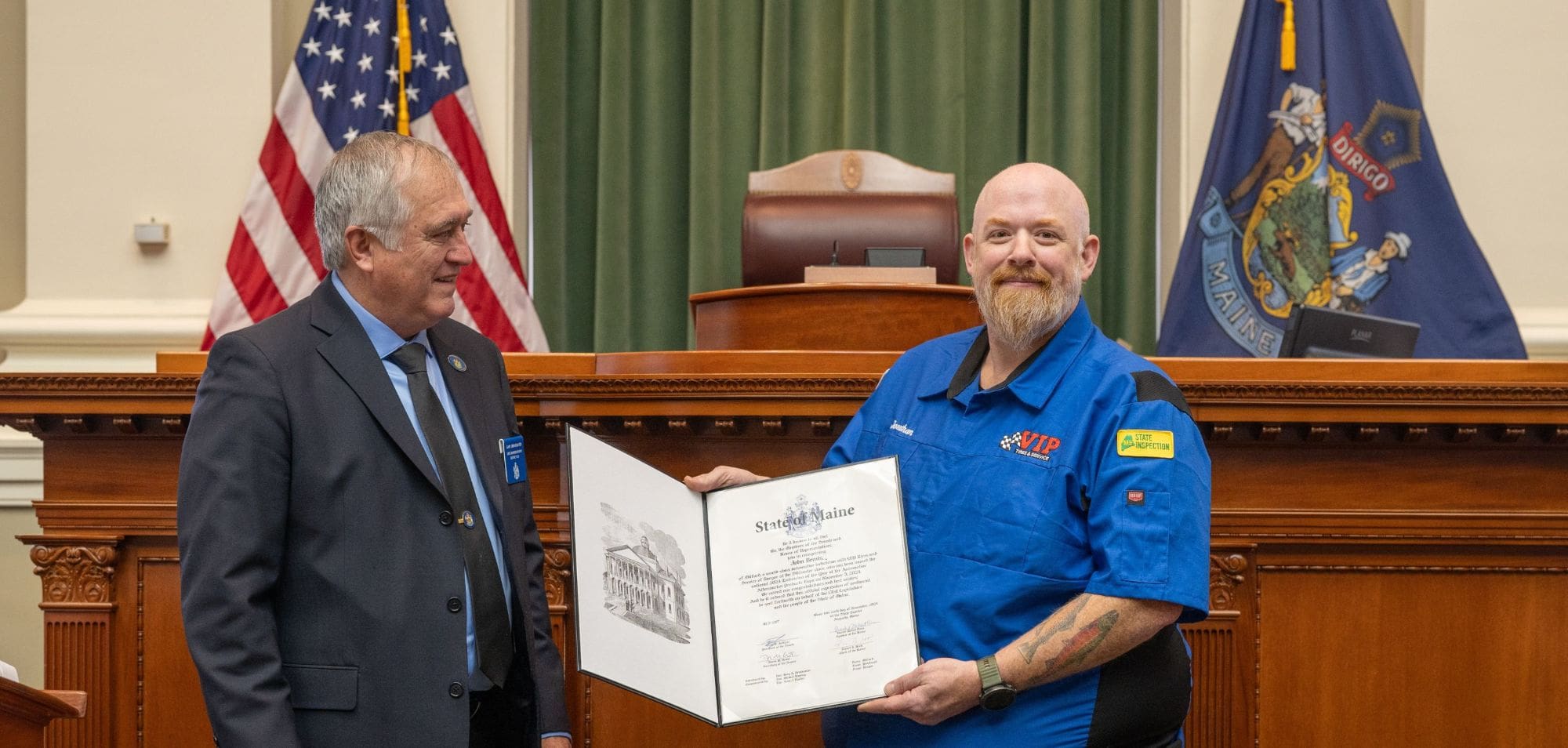 Two men hold an open certificate in front of an American flag and state flag in an official.